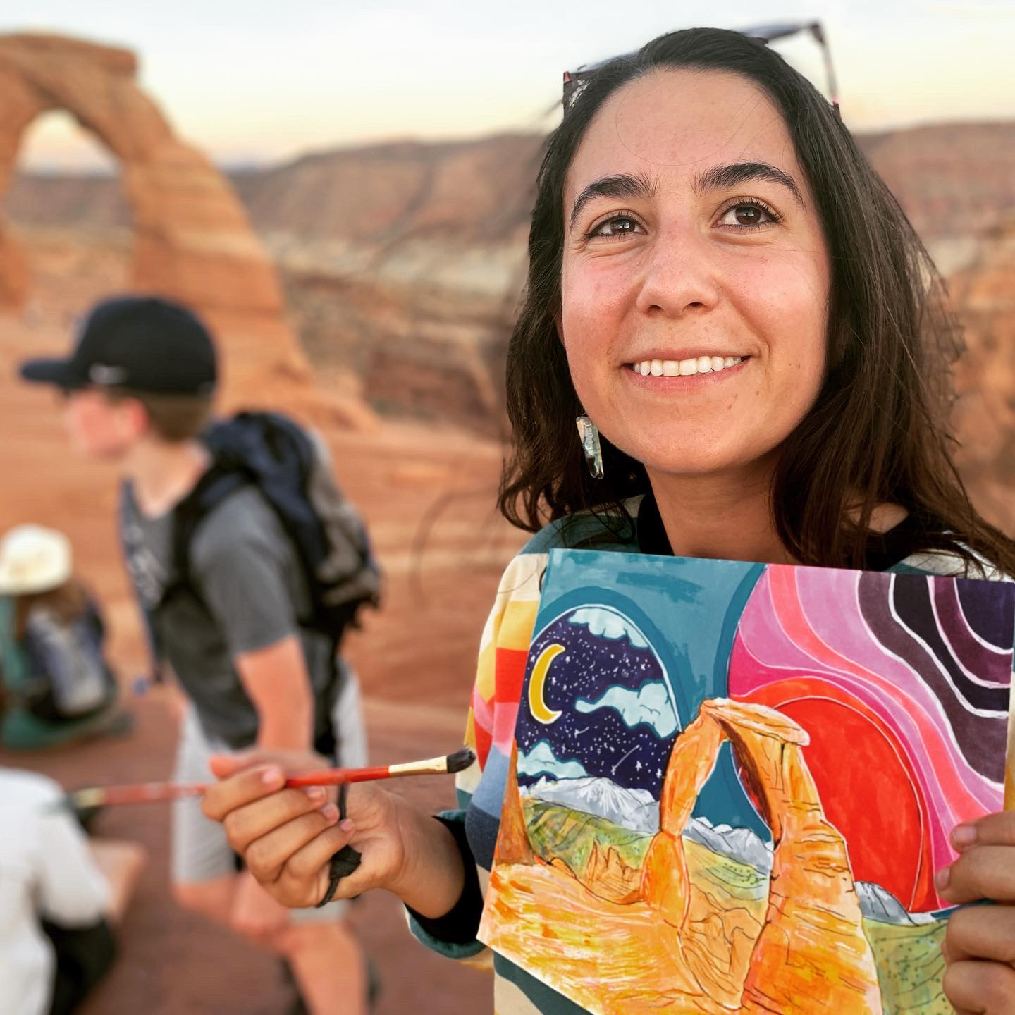 Alina holds a painting off a groovy colourful depiction of Delicate Arch while standing in front of the real thing.
