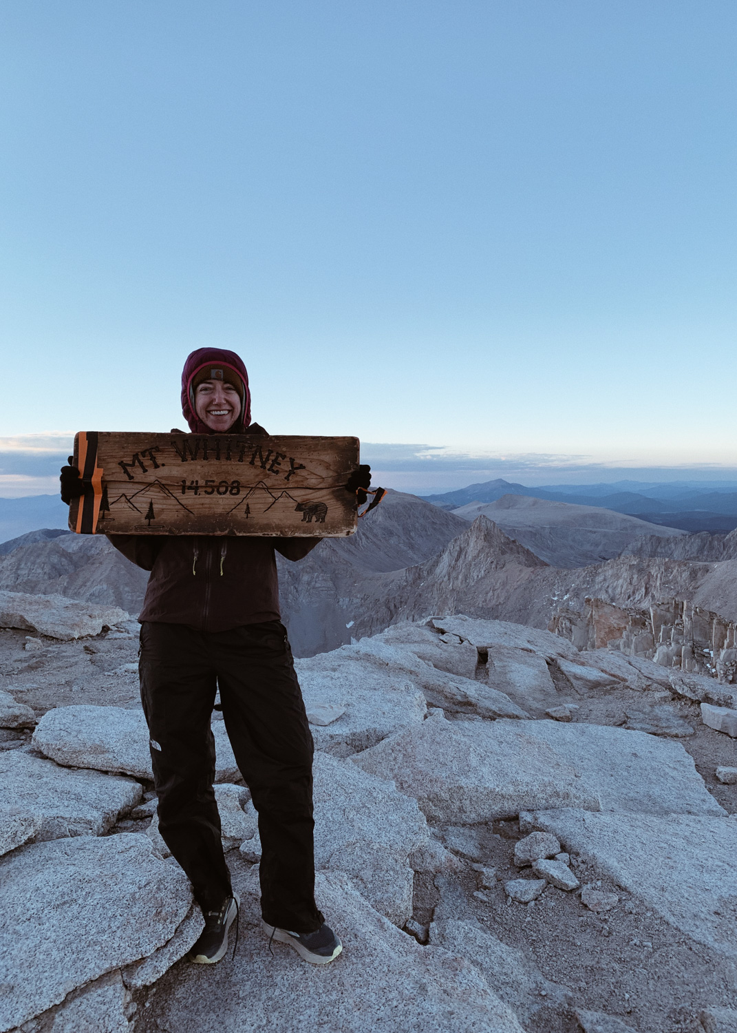 A female hiker smiles atop Mt Whitney (Tumanguya) with a wooden sign just before sunrise